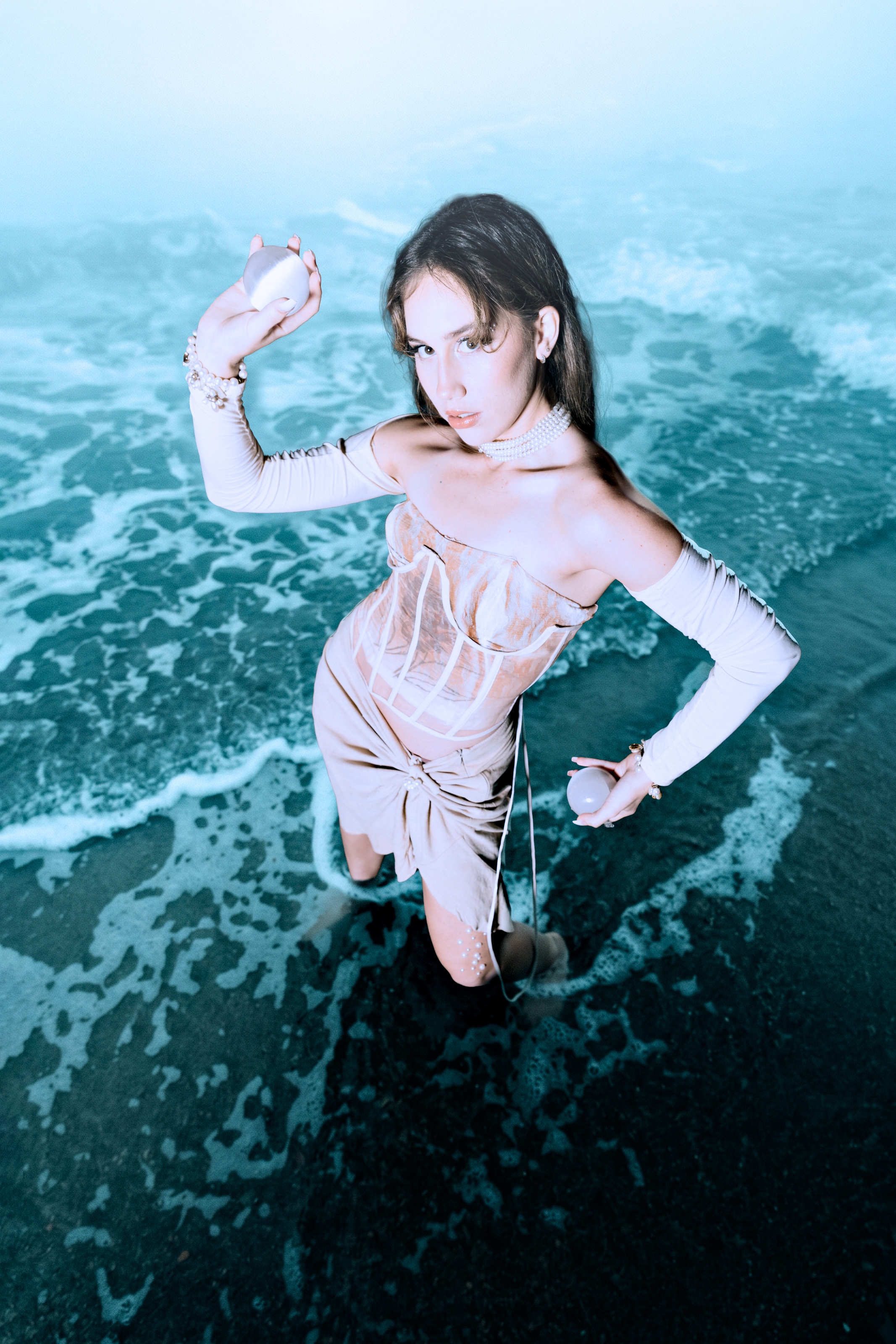 Sığ deniz suyunda duran ve inciyi andıran iki küre tutan bir modelin yukarıdan çekilmiş portresi. Fildişi giysiler, inci aksesuarlar ve kıvrılan deniz köpüğü çarpıcı bir editoryal kompozisyon oluşturuyor.